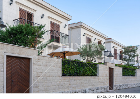 A row of elegant modern villas with balconies and lush greenery, enclosed by a stone wall, showcasing upscale residential living. 120494010