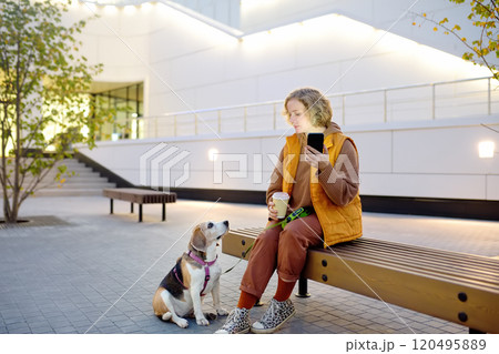 Hipster woman drinks coffee while walking with old Beagle dog along city street. Girl, dog owner, searches for pet products on the Internet using smartphone applications. Vet emergency care for pets 120495889
