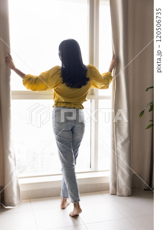 Back view of woman parting curtains at large window 120506735