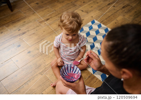 A mother lovingly feeds her curious toddler with a spoonful of pink yogurt while they sit on a wooden floor. 120509694