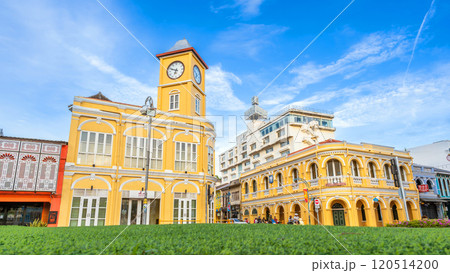 The Sino-Portuguese yellow clock tower and Chartered Bank building in Phuket Old Town, surrounded by lush greenery, stand out under a vivid blue sky, showcasing colonial charm 120514200