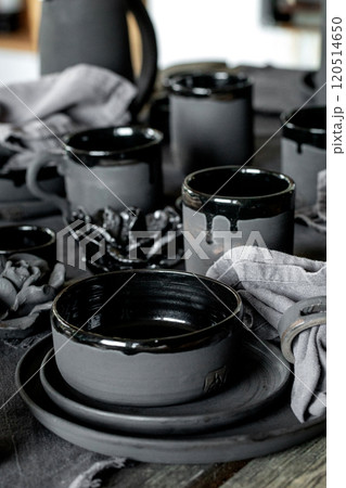 Rustic table setting with empty craft handmade ceramic tableware, black bowls, plates, cups, jug and vase on linen tablecloth. Blck ceramic roses flowers decor. Day light dining room. Rustic table setting with empty craft handmade ceramic tableware, black bowls, plates, cups, jug and vase on linen tablecloth. Blck ceramic roses flowers decor. Day light dining room. 120514650