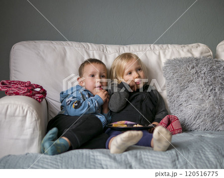 Two kids sit on white couch, enjoying snacks and watching tv under cozy blanket with fluffy pillows. The relaxing atmosphere creates warm moment as two kids unwind together comfortably. 120516775