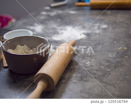 Wooden rolling pin sits on a cluttered, flour-dusted kitchen counter alongside a bowl of flour, hinting at the making of cookie dough 120516784