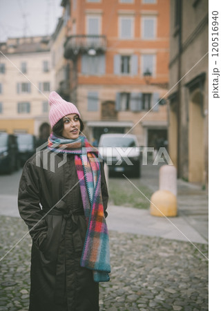 A woman in a pink hat and scarf stands on a cobblestone street 120516940
