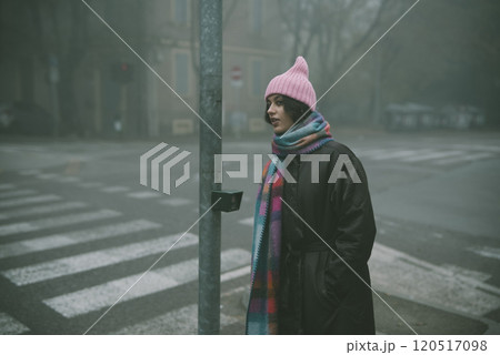 A woman, wearing a pink hat and scarf, stands on a foggy street 120517098