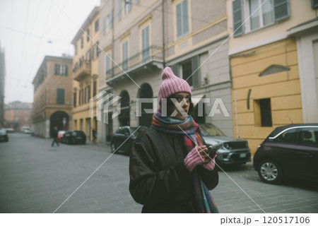 A woman in a pink hat and scarf stands on a street, looking at her phone 120517106