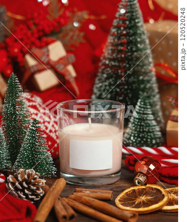 Glass candle with label near red knitted sweater and Christmas decor and lights, winter mockup 120520248