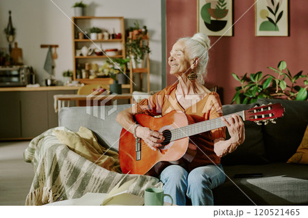 Senior woman happily playing guitar while relaxing on sofa with cozy surroundings in background including shelves, plants, and home decor creating an intimate, homely atmosphere 120521465