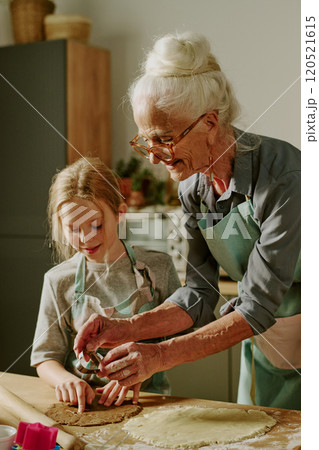 Elderly woman teaching young girl baking, both enjoying shared activity in cozy kitchen setting. Senior and child wearing matching aprons, surrounded by kitchen tools and ingredients Elderly woman teaching young girl baking, both enjoying shared activity in cozy kitchen setting. Senior and child wearing matching aprons, surrounded by kitchen tools and ingredients 120521615