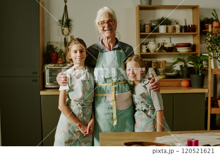 Senior woman baking with two young girls, wearing aprons and smiling together in kitchen, standing near wooden table with baking supplies 120521621