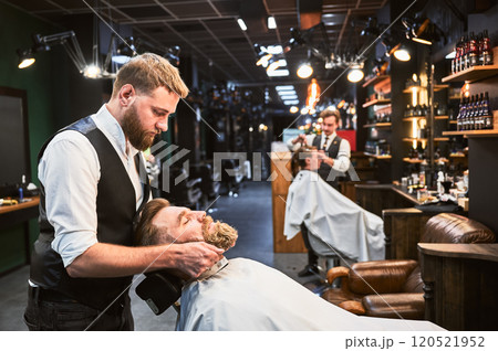 Professional barber shaving client's beard. Modern barbershop with fancy interior. Hairdresser in elegant outfit at work. 120521952