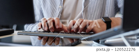 Close-up image showing persons hands typing on keyboard, with reflections of fingers and screens visible. Modern gadgets and workplace environment evident in background 120522276