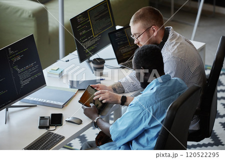 Two colleagues working together on software development in modern tech workspace, both focused on coding and using digital tools. One person is showing information on a tablet Two colleagues working together on software development in modern tech workspace, both focused on coding and using digital tools. One person is showing information on a tablet 120522295