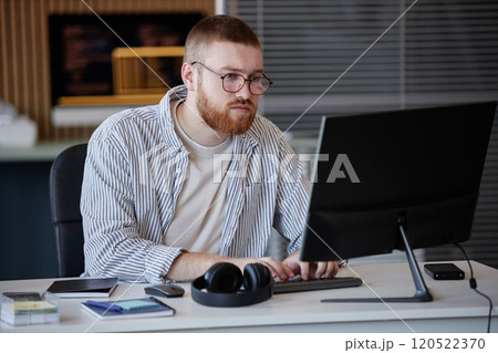 Man with glasses sitting at desk engaging with computer in spacious office environment. Various items such as phone, headphones, and notepads placed around working area Man with glasses sitting at desk engaging with computer in spacious office environment. Various items such as phone, headphones, and notepads placed around working area 120522370