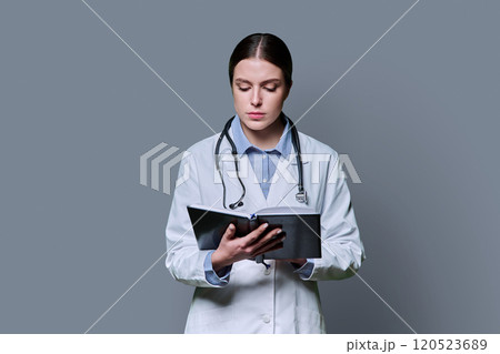 Portrait of young female doctor looking at notebook on gray background 120523689