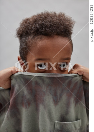 Vertical shot of funny little Black boy hiding face in t shirt pulling up collar and peeking out at camera, while standing against gray background in studio 120524255