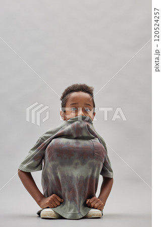 Vertical shot of funny little Black boy sitting on squats pulling t shirt on knees while peeking out from clothes playfully staring at camera against grey background in studio, copy space Vertical shot of funny little Black boy sitting on squats pulling t shirt on knees while peeking out from clothes playfully staring at camera against grey background in studio, copy space 120524257