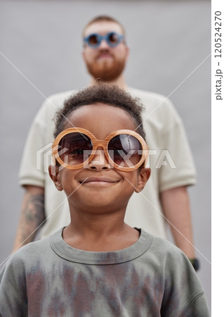 Vertical portrait shot of cheerful little Black boy in large sunglasses happily smiling, while adoptive father standing in background wearing resembling accessory against gray background in studio Vertical portrait shot of cheerful little Black boy in large sunglasses happily smiling, while adoptive father standing in background wearing resembling accessory against gray background in studio 120524270