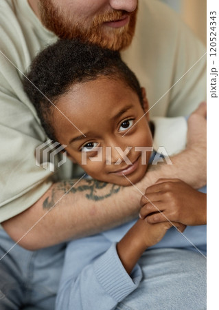 Vertical portrait shot of little smiling Black boy in tender embrace of adoptive father happily looking at camera while spending time with parent at home 120524343