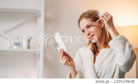 Beauty care. Female routine. Pleasant smiling lady applying makeup using powder brush looking in mirror enjoying her reflection in white robe while holding cosmetic tools in hand. Beauty care. Female routine. Pleasant smiling lady applying makeup using powder brush looking in mirror enjoying her reflection in white robe while holding cosmetic tools in hand. 120529626
