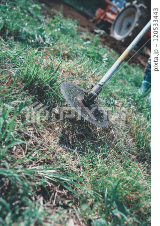 畑周りの雑草を刈払機で刈る女性 120533443