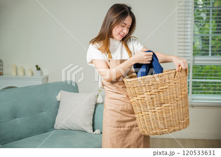 Asian housewife woman holding basket with heap of different clothes on sofa in living room, Beautiful housekeeper doing housework holds wooden basket of clean clothes to washing at home Asian housewife woman holding basket with heap of different clothes on sofa in living room, Beautiful housekeeper doing housework holds wooden basket of clean clothes to washing at home 120533631