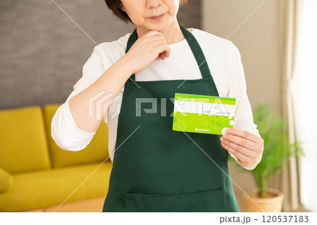 家のリビングで通帳を持って悩む緑のエプロンをしたミドル〜シニア女性の主婦 120537183