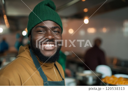 A smiling man in a green beanie and apron stands in a kitchen, ready to serve food to others 120538313
