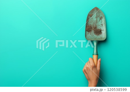 A hand gripping a dirt covered trowel with a wooden handle on a turquoise background 120538599