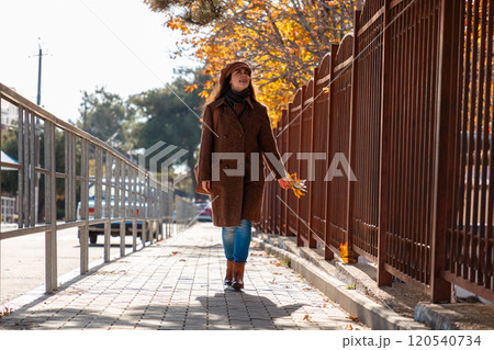 Wide shot of young pretty Caucasian woman in a coat holds bunch of yellow leaves and walking outdoors in city street on autumn day. Lifestyle and city life Wide shot of young pretty Caucasian woman in a coat holds bunch of yellow leaves and walking outdoors in city street on autumn day. Lifestyle and city life 120540734