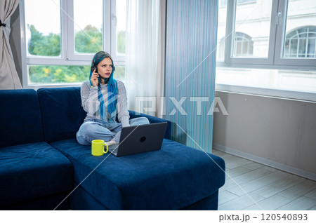 Beautiful blue haired hipster woman is working from home sitting on her couch, wearing headphones with microphone to attend a meeting with laptop. Concept of working from home, using technology. Beautiful blue haired hipster woman is working from home sitting on her couch, wearing headphones with microphone to attend a meeting with laptop. Concept of working from home, using technology. 120540893
