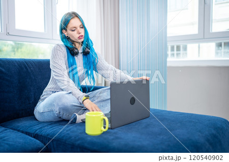Beautiful blue haired hipster woman is working from home sitting on her couch, wearing headphones with microphone to attend a meeting with laptop. Concept of working from home, using technology. Beautiful blue haired hipster woman is working from home sitting on her couch, wearing headphones with microphone to attend a meeting with laptop. Concept of working from home, using technology. 120540902