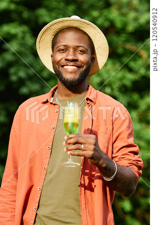 Smiling African American man wearing a hat and necklace while holding a drink and standing outdoors in natural surroundings, exuding a joyful and relaxed vibe Smiling African American man wearing a hat and necklace while holding a drink and standing outdoors in natural surroundings, exuding a joyful and relaxed vibe 120540912