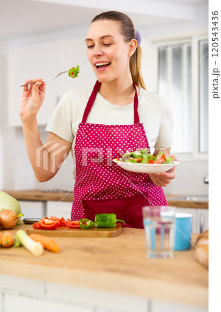 Positive housewife eating homemade salad in kitchen 120543436