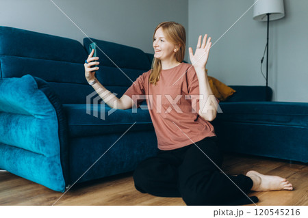 Hi. Female taking a selfie photo using her mobile phone at home. woman in t-shirt recording video, talking on online call and relaxing on floor near sofa. Communication and comfort concept, full body Hi. Female taking a selfie photo using her mobile phone at home. woman in t-shirt recording video, talking on online call and relaxing on floor near sofa. Communication and comfort concept, full body 120545216