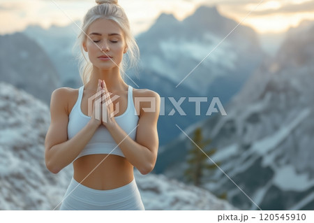 Blonde woman with hands in prayer pose, standing on a mountain with snow covered peaks behind her 120545910