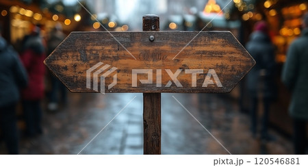 A rustic arrow shaped wooden sign stands in the middle of a festive market, with blurred lights behind 120546881