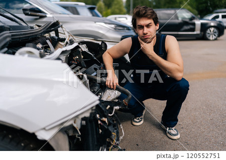 Thoughtful mechanic male in uniform examining front of car without bumper, assessing plan for future work in automobile repair and renew service station. Concept of car service, repair, maintenance. Thoughtful mechanic male in uniform examining front of car without bumper, assessing plan for future work in automobile repair and renew service station. Concept of car service, repair, maintenance. 120552751