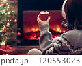 Woman in headphones with cup of hot cocoa and marshmallow sitting and warming at winter evening near fireplace flame and  christmas tree. 120553023