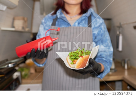 Medium close up of unrecognizable hands of female cook holding hot dog and ketchup squeeze bottle Medium close up of unrecognizable hands of female cook holding hot dog and ketchup squeeze bottle 120554373