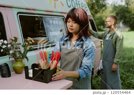 Medium close up of Asian young woman with stripped apron adjusting plastic box with multicolored straws and paper tissues on table next to food truck Medium close up of Asian young woman with stripped apron adjusting plastic box with multicolored straws and paper tissues on table next to food truck 120554464