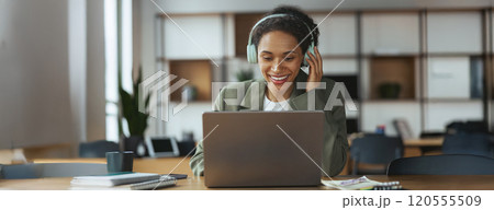 A woman with headphones smiles at her laptop on the table in nice modern coworking office 120555509