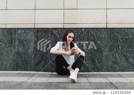 Young woman sitting on pavement while using smartphone in urban setting 120556304