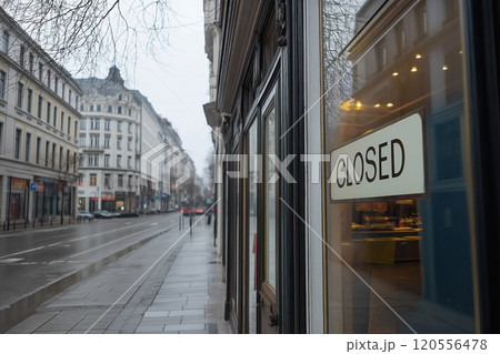 A closed sign reflected on a glass window of a shop, with a quiet, empty city street outside 120556478