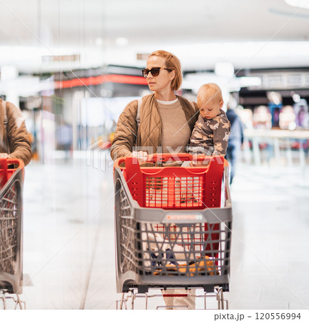 Mother shopping with his kid in the mall. Mother pushing shopping cart while holding and carrying her toddler baby boy child along mall corridor. Mother shopping with his kid in the mall. Mother pushing shopping cart while holding and carrying her toddler baby boy child along mall corridor. 120556994