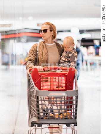 Mother shopping with his kid in the mall. Mother pushing shopping cart while holding and carrying her toddler baby boy child along mall corridor. 120556995