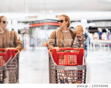 Mother shopping with his kid in the mall. Mother pushing shopping cart while holding and carrying her toddler baby boy child along mall corridor. Mother shopping with his kid in the mall. Mother pushing shopping cart while holding and carrying her toddler baby boy child along mall corridor. 120556996
