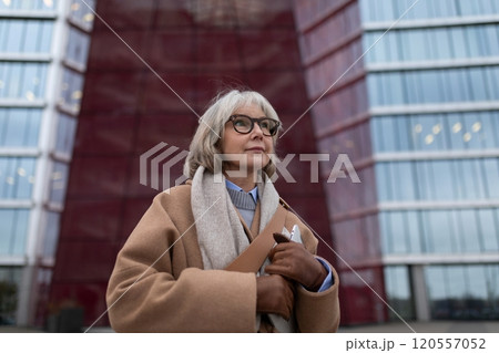 An elderly woman with glasses poses confidently in front of a modern building with glass and red 120557052