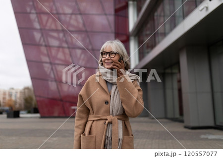 A smiling elderly woman with glasses enjoys a stroll outside a modern building during a cloudy day 120557077